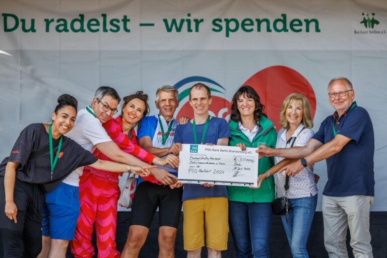 Eine Gruppe von Menschen hält gemeinsam einen großen Scheck für eine Spendenaktion, lächelnd vor einem Banner mit der Aufschrift „Du radelst – wir spenden“.